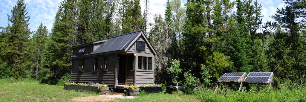 A small transportable home in a forest clearing, with two solar panels on a ground frame beside it