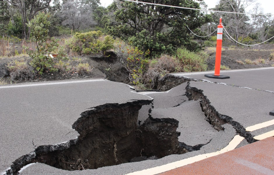 A road surface split open and collapsed after an earthquake, with a traffic cone beside the hole