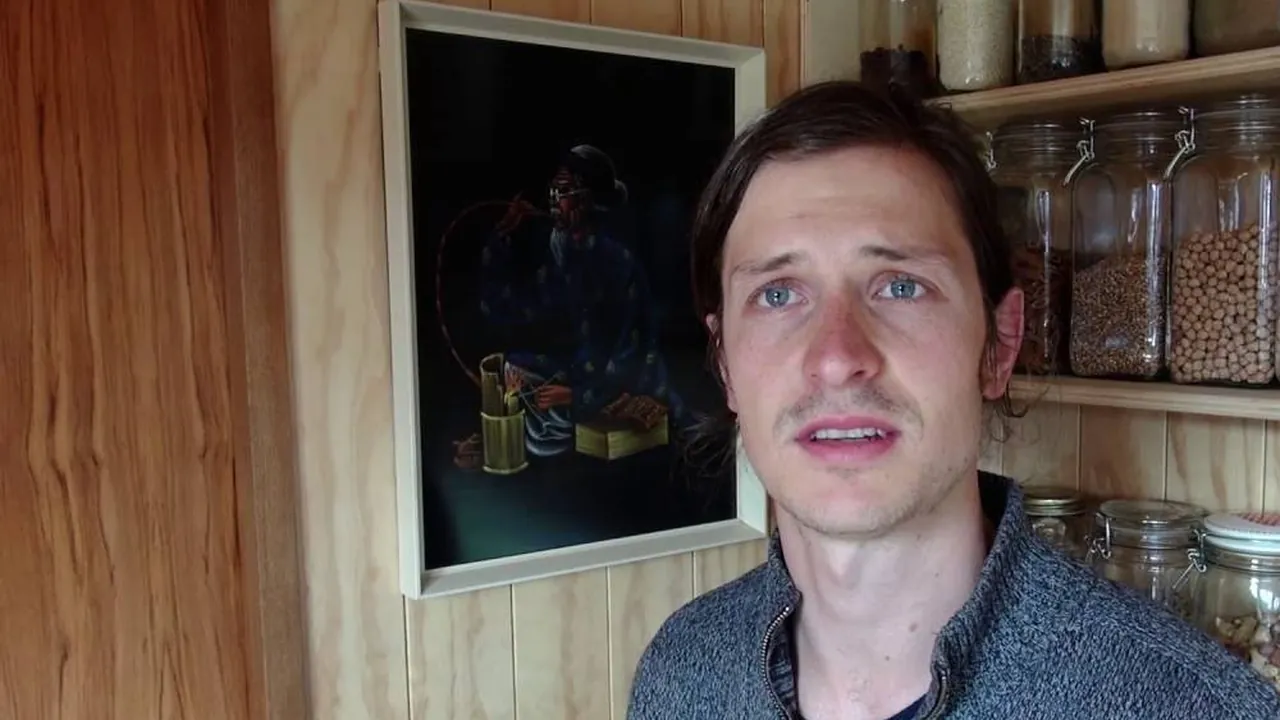 Kyle Sutherland talking to the camera in his tiny house, in front of shelves of glass storage jars